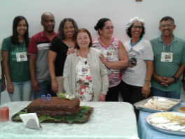 Cinco mulheres e dois homens em frente a um bolo de chocolate, que conta com uma vela sinalizando 20 anos.