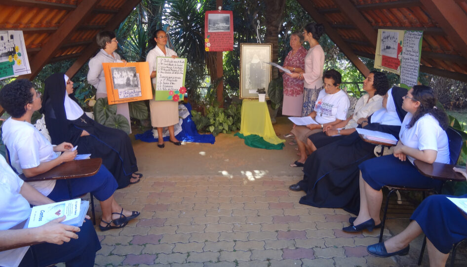 Grupo de mulheres em roda, assistindo uma apresentação feita por quatro delas, que seguram cartazes.