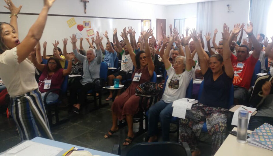 Grupo de pessoas em uma sala de aula, levantando as mãos para o alto.