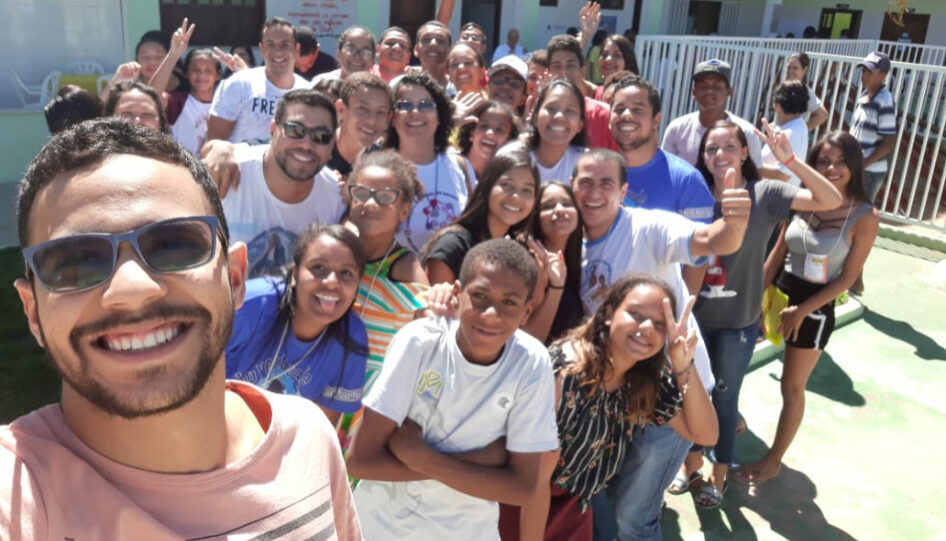 Um homem tirando uma selfie com um grupo de pessoas, a maioria crianças, em um ambiente que se assemelha a um pátio de uma escola.